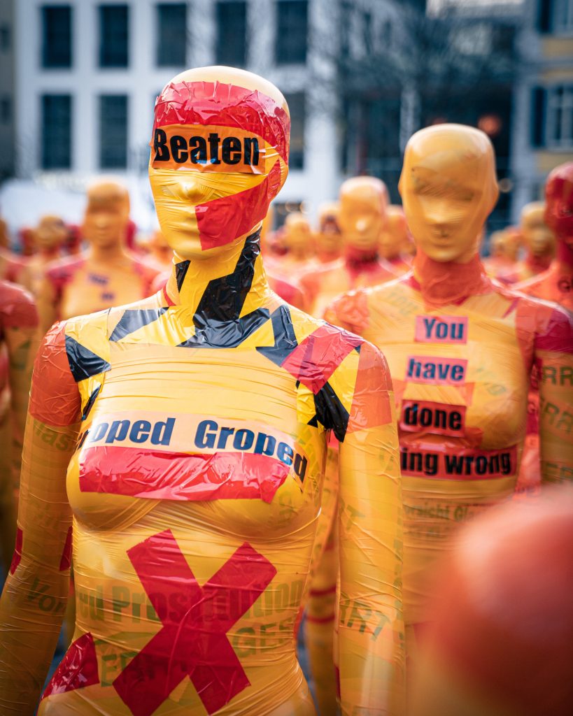 A mannequin wrapped in yellow tape with "beaten" written over its eyes and "groped groped" written on its chest. There is another mannequin with "you have done nothing wrong" written on its chest.