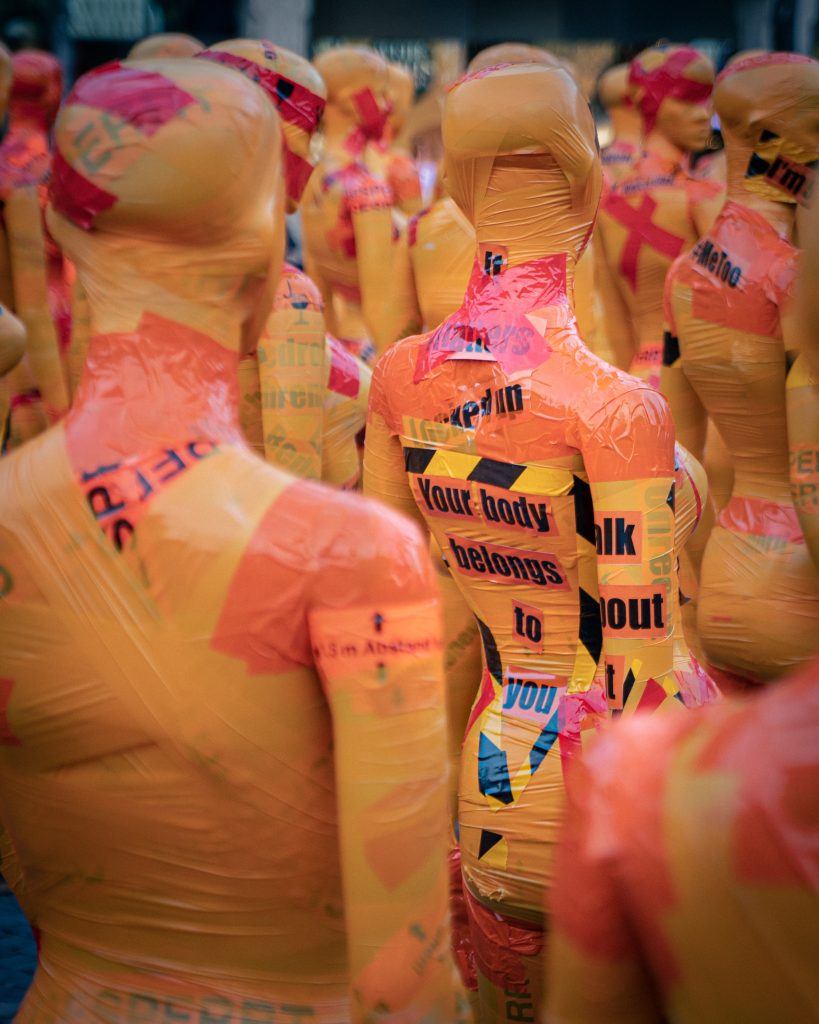 A multitude of mannequins wrapped in yellow and red tape. One mannequin has "Your body belongs to you" written on its back.
