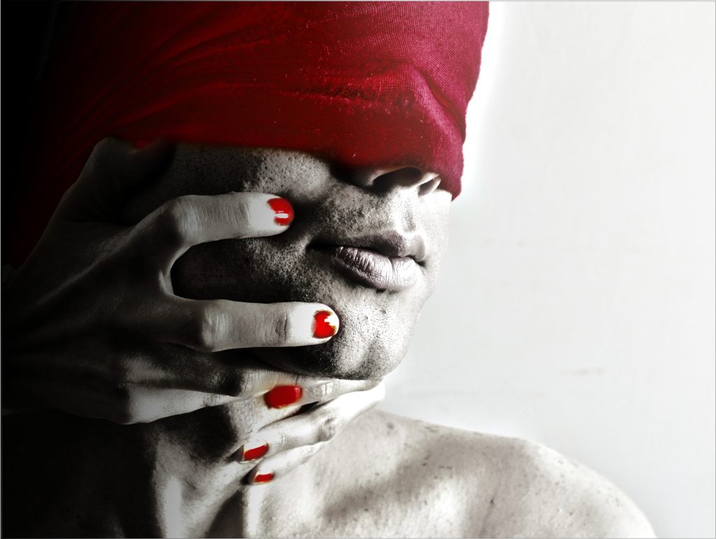 Hands with red painted nails grip the jaw of someone with a red blindfold on. 