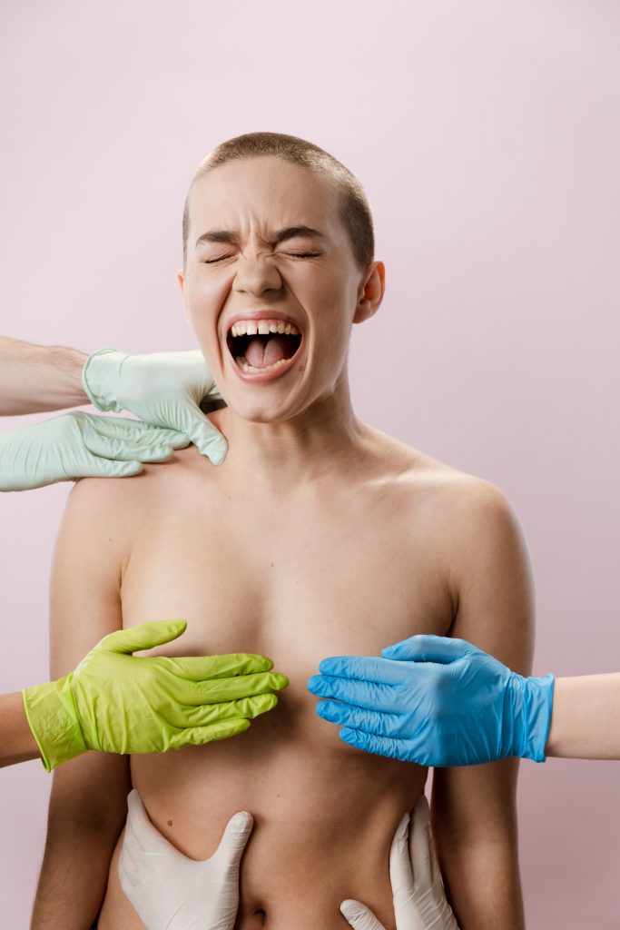 A person is standing shirtless with gloved hands touching their body. They have their mouth open as if they are yelling.