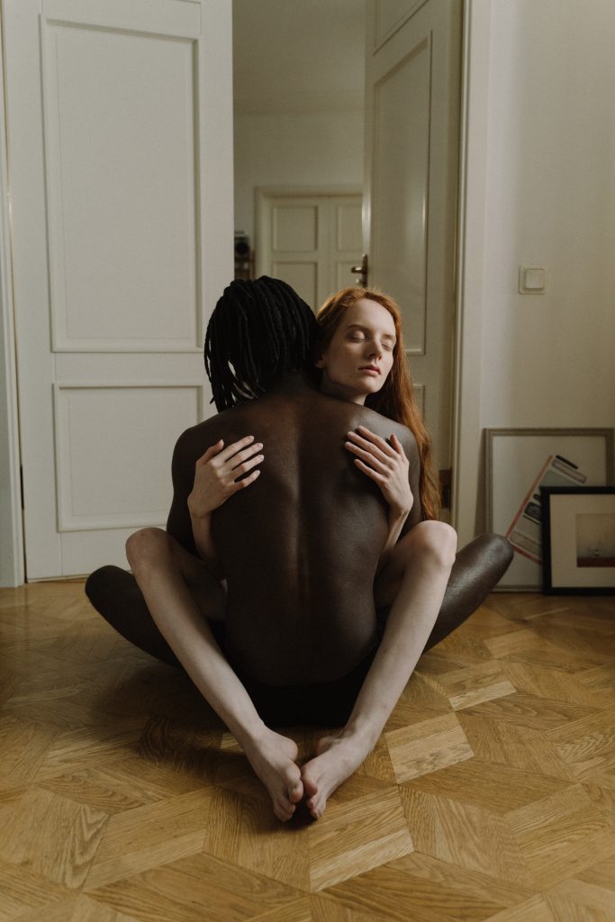 A nude couple hugging while sitting on the floor.