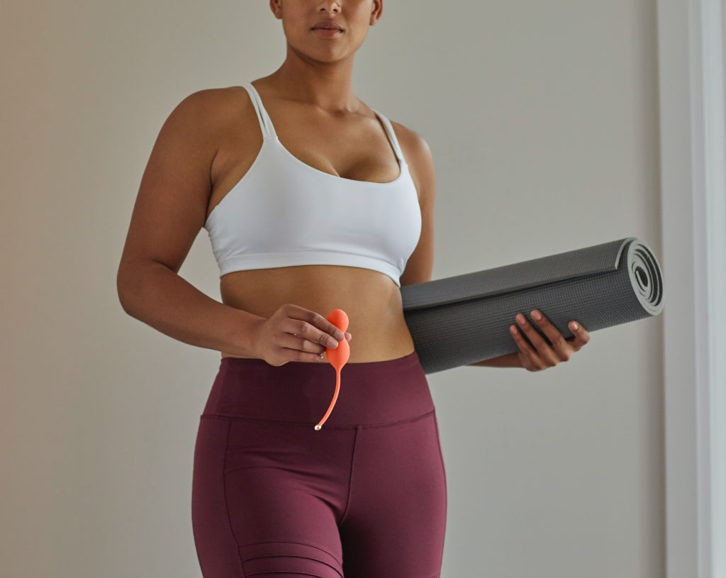 A woman in exercise clothes holding Kegel balls and a yoga mat.