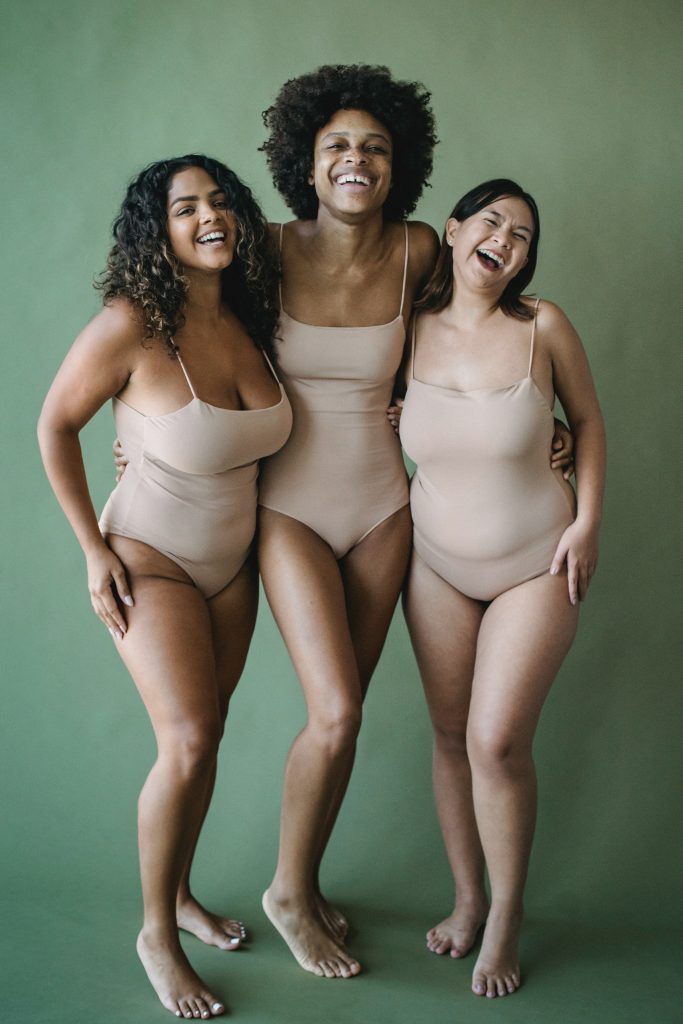 Three individuals in tan bodysuits. They are smiling and hugging one another.