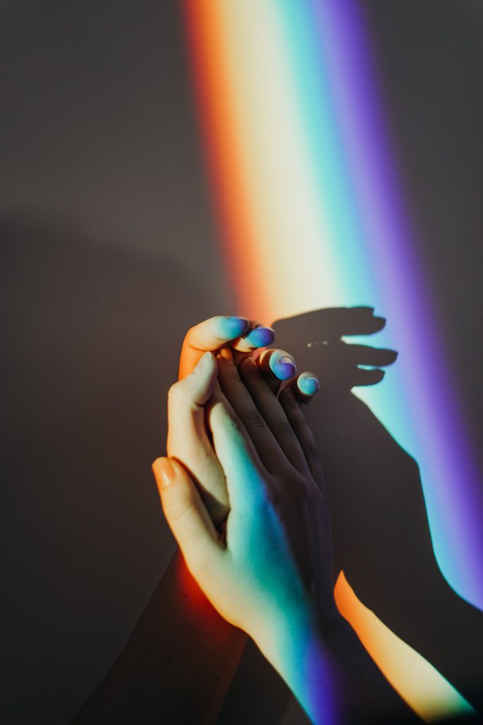 A pair of hands holding each other. There is a rainbow light hovering over the hands.