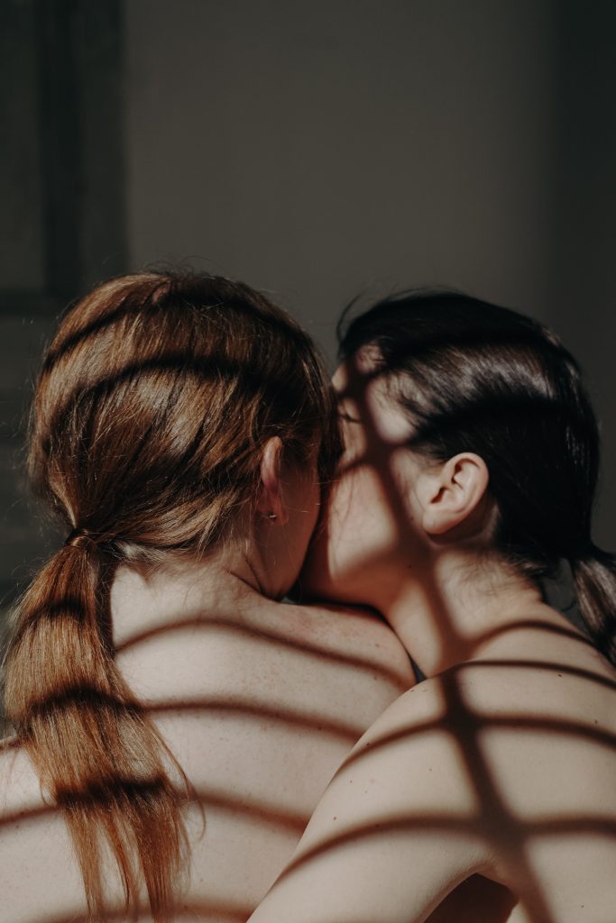 A topless couple facing their backs to the camera. One person is kissing the other's cheek.