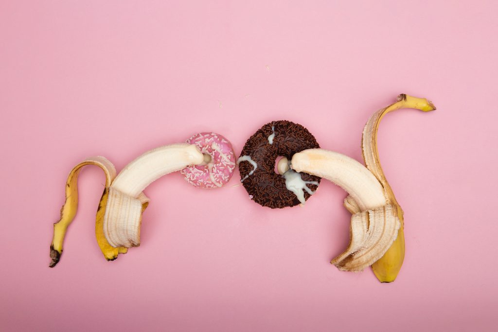 One banana entering a pink sprinkles donut and another banana entering a chocolate sprinkles donut. There is a milky substance on the donuts.