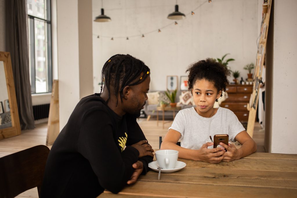 An adult and a child sitting a table. They are looking at each other and the child is holding a phone.