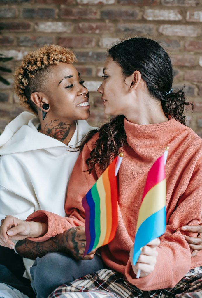 Two individuals looking into each other's eyes. One individual is holding an LGBTQ flag and a pansexual flag.