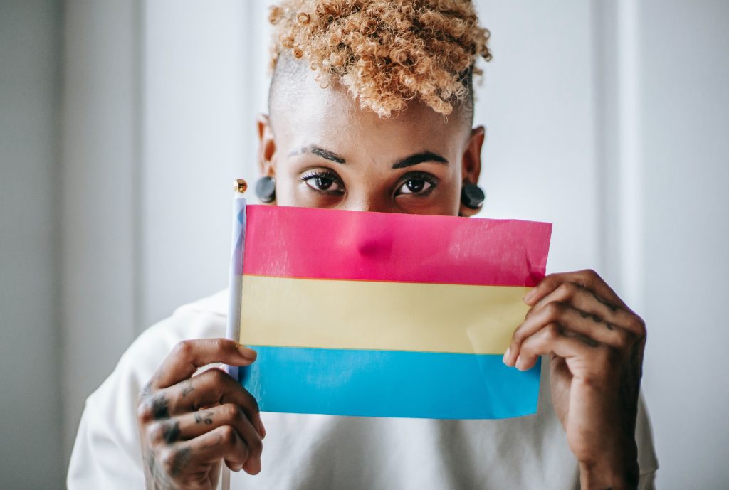 A person holding a bisexual flag over their face. The flag is three horizontal blocks; the top color is red, the middle is yellow, and the bottom is blue.