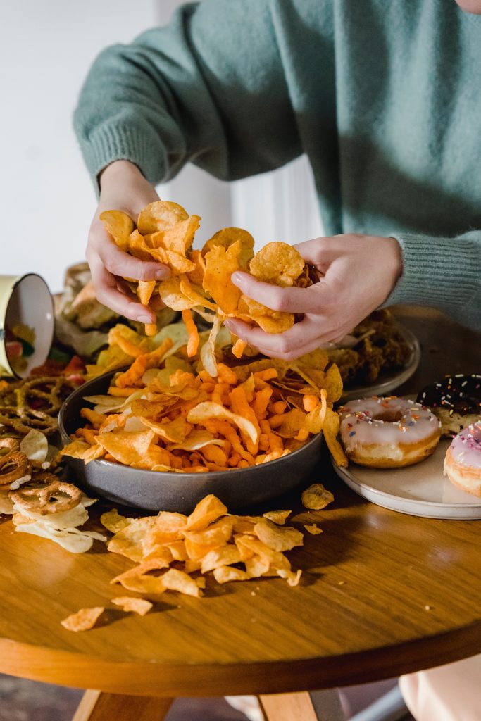 Chips, donuts, and pretzels on a table. A person is holding two handfuls of chips.