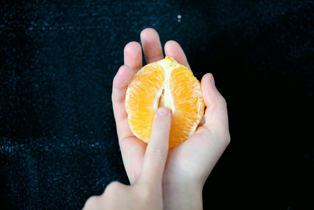 A person's finger over a half-cut orange that resembles a vagina.