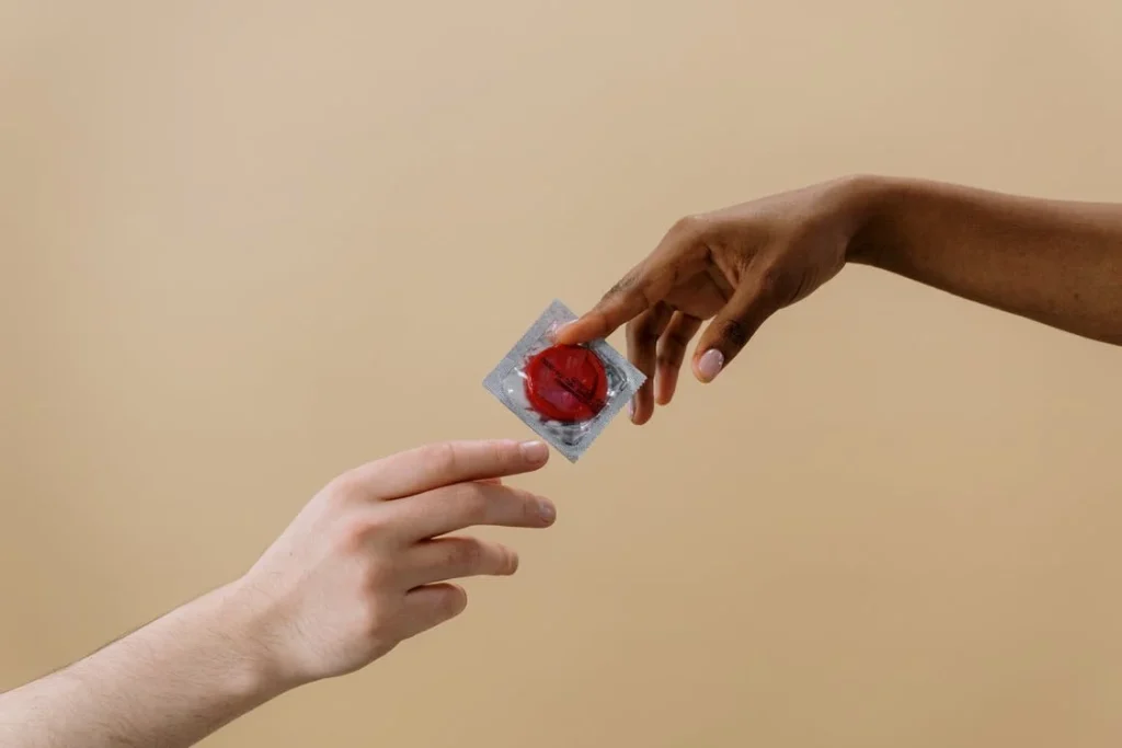 two hands reaching towards each other; one is holding a sealed condom package with a red condom inside, and the other hand is reaching out with its fingers towards the condom package. 
