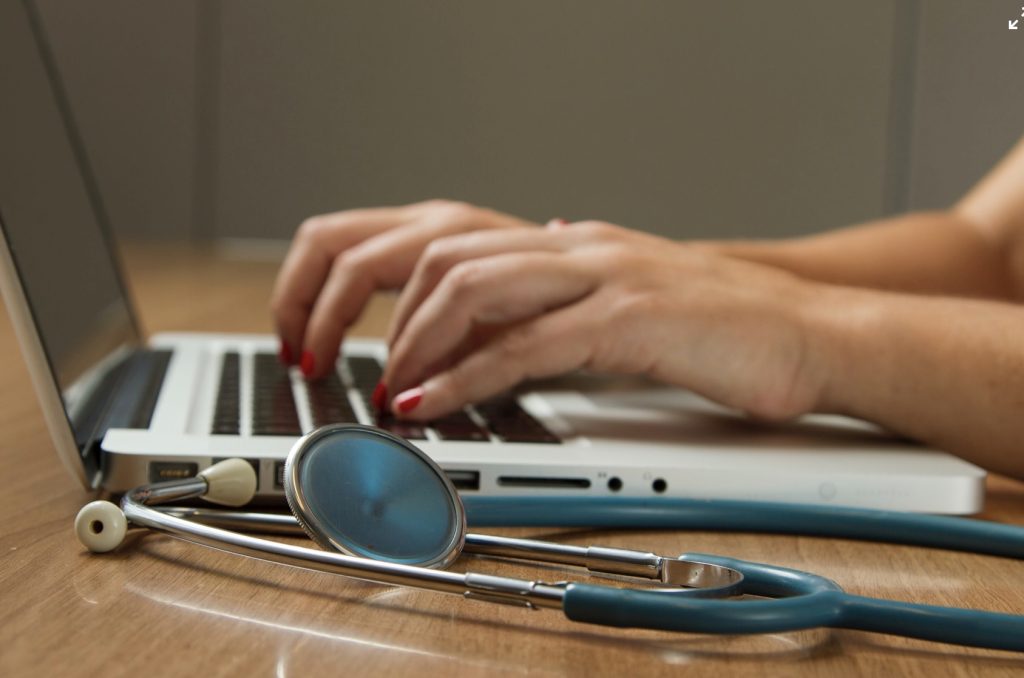 hands typing on computer on brown desk, blue stethoscope next to computer
