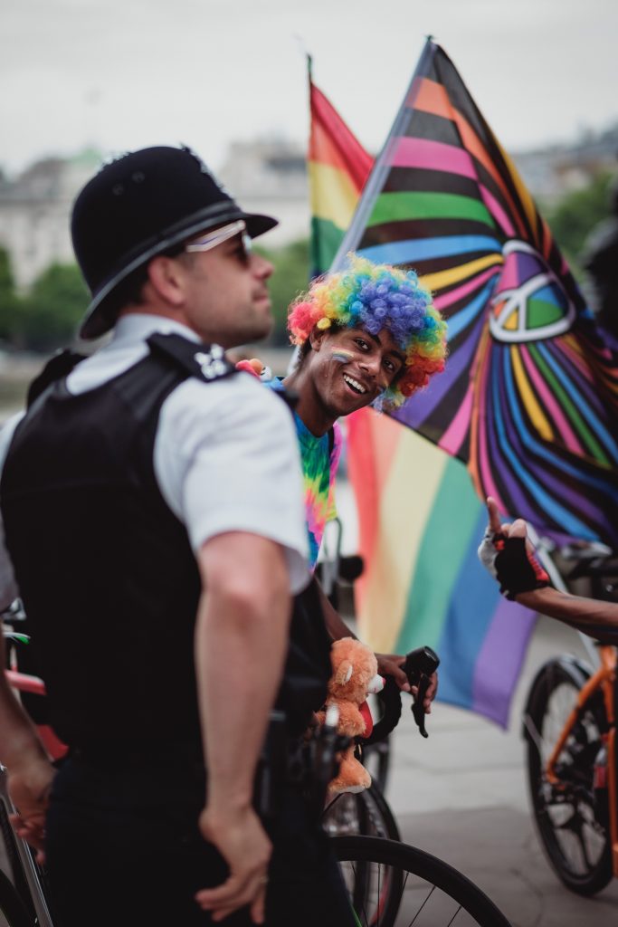 Man dressed in all rainbow colors gives the camera a smile at a pride event from behind a police officer who is looking in a different direction