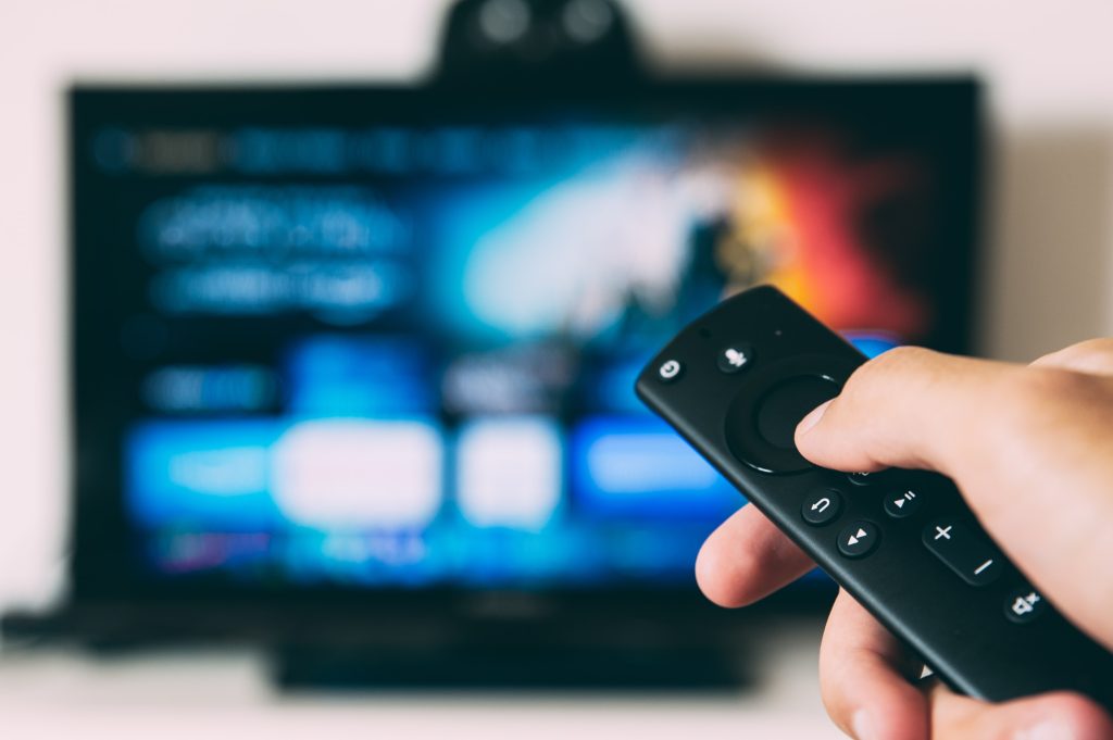 A hand is holding a remote control with a blurry TV in the background. 