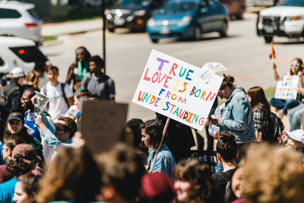 Multiple people protesting outdoors. There is one sign with multicolored writing that spells "true love is born from understanding."