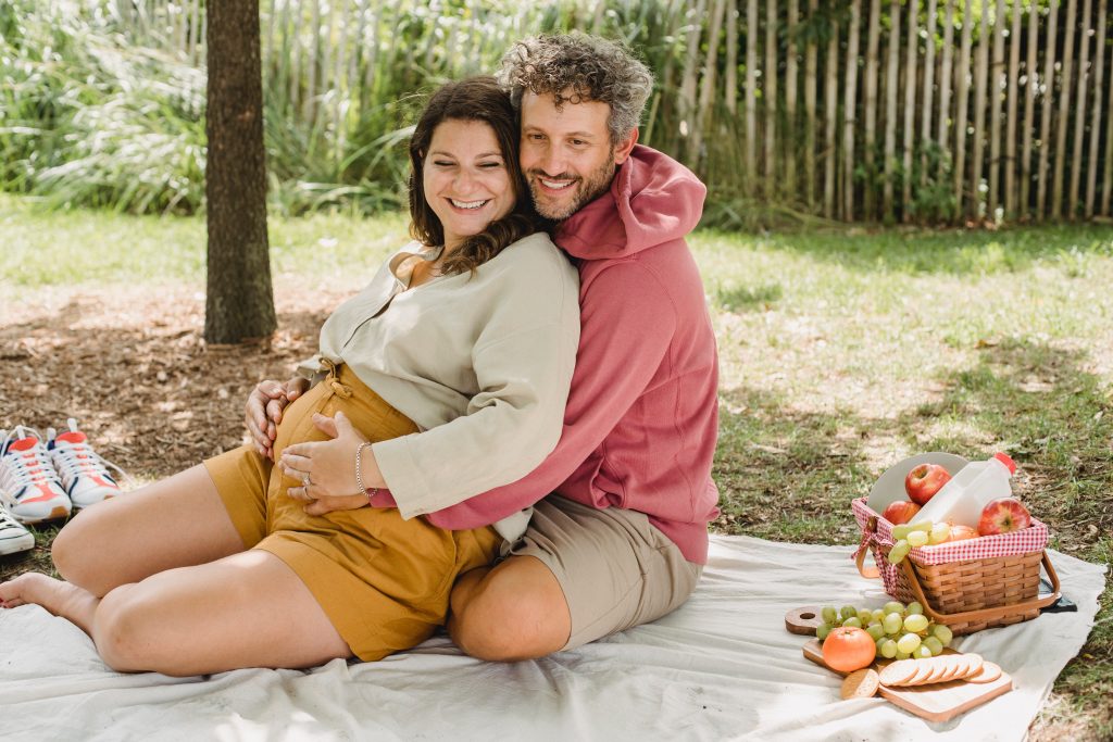 A man hugging a pregnant woman while they sit on a sheet on top of grass. There is a picnic basket filled with fruit beside the couple.