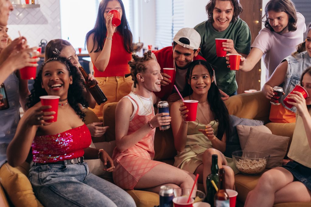 A group of individuals sitting around a couch. They are all smiling and holding red solo cups.