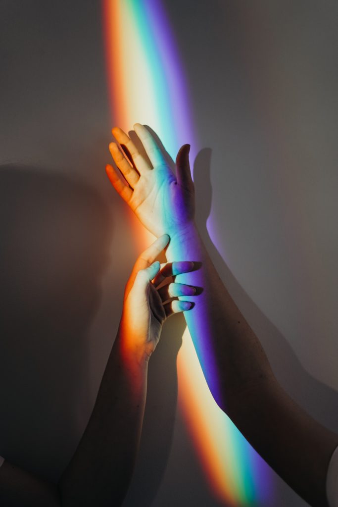 A couple hands reaching upward. There is a rainbow shining on them.