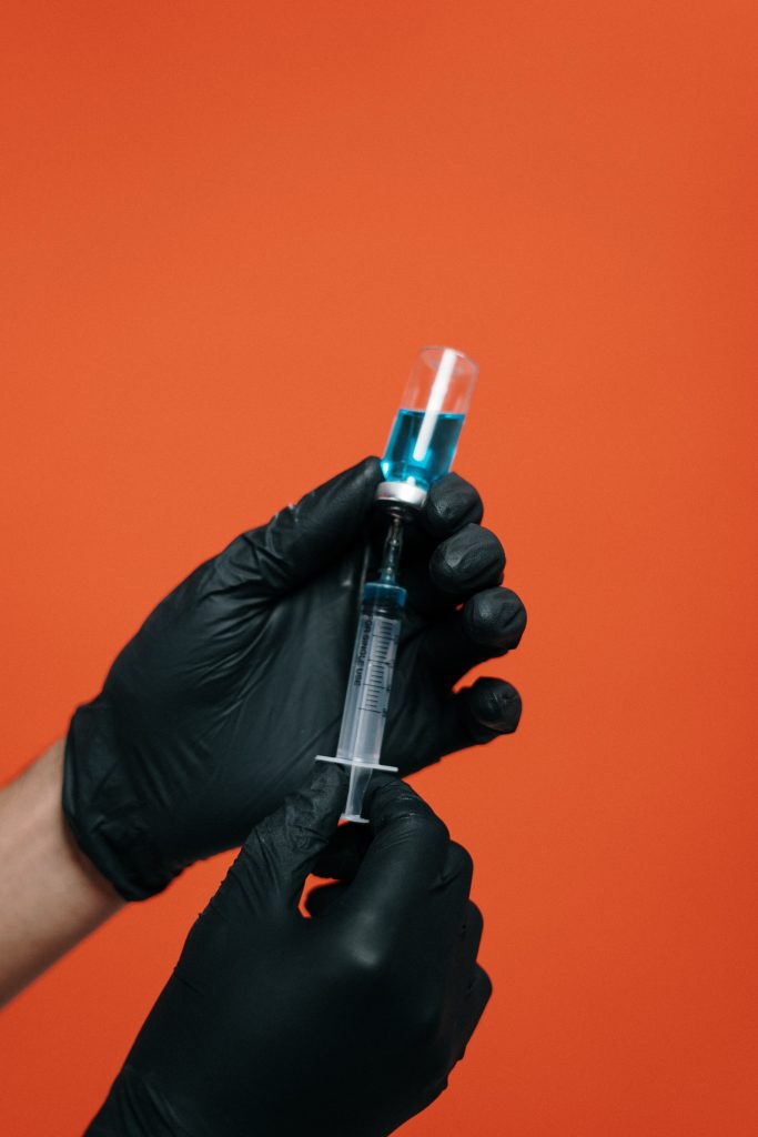 A pair of black gloved hands drawing up a vaccine with a syringe. 
