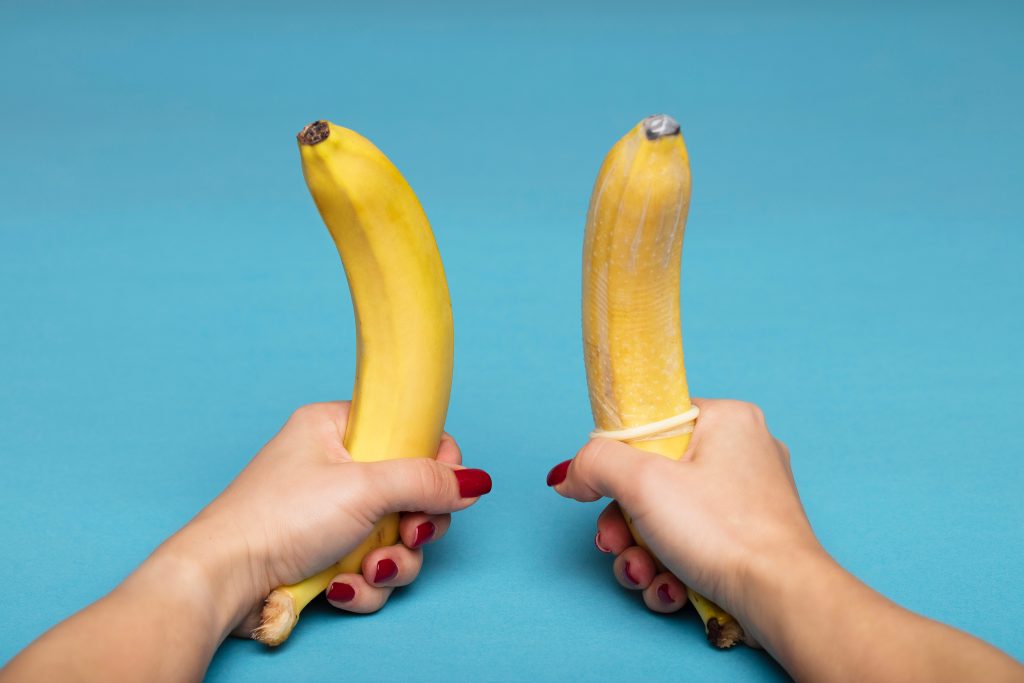 Two bananas being held by hands.. The banana on the right has a condom on it.