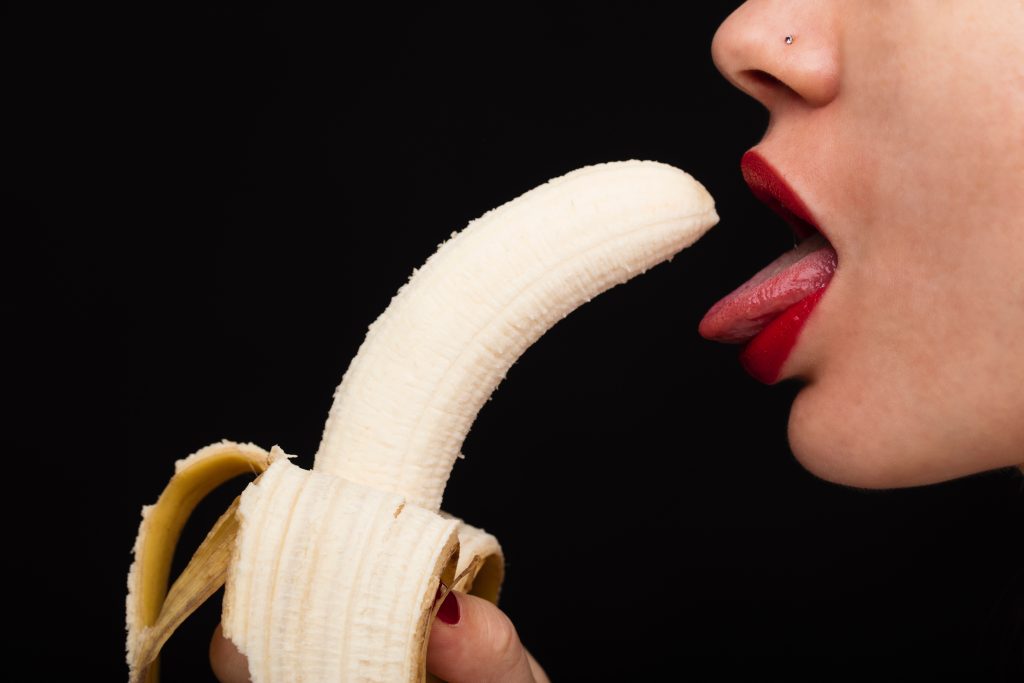 A person's tongue coming out of their mouth and toward a peeled banana.
