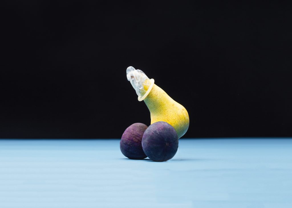 A pair with a fig to its right and a fig to its left. There is a condom on the tip of the pear. The pear resembles a penis and the figs resemble testicles.