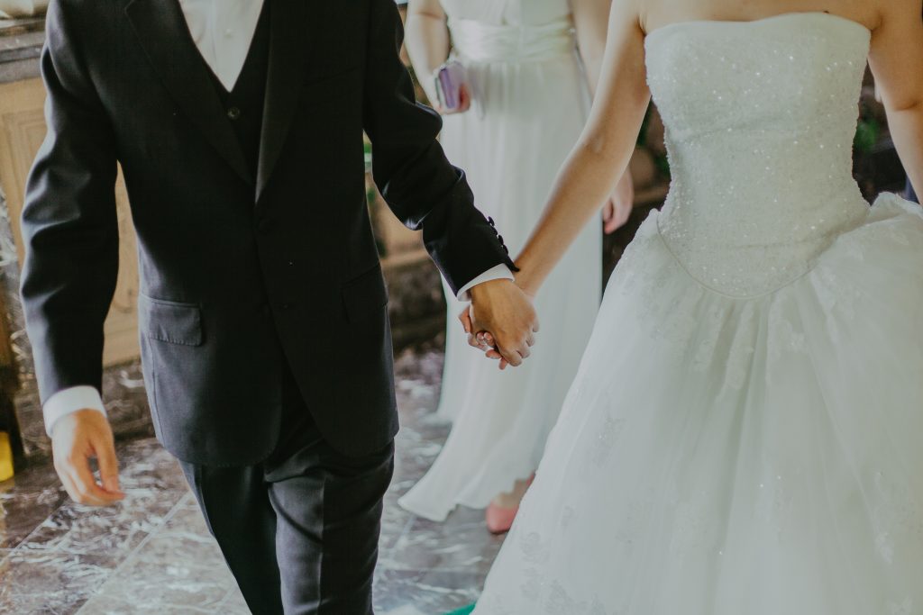 An individual in a suit holding hands with an individual in a white wedding dress.