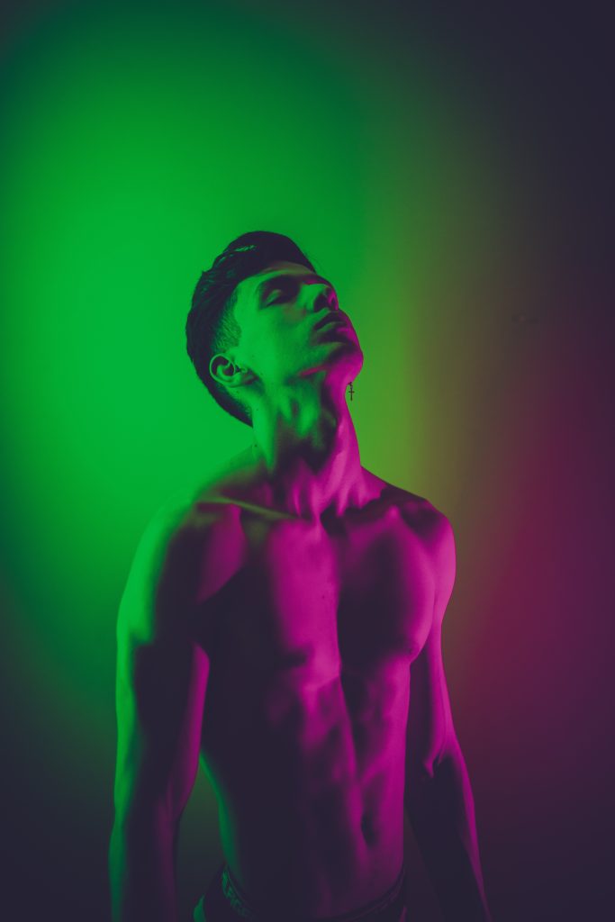 A topless man with pink and green light reflecting on him.