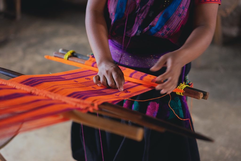 Native American person weaving a bright orange and purple colored cloth.