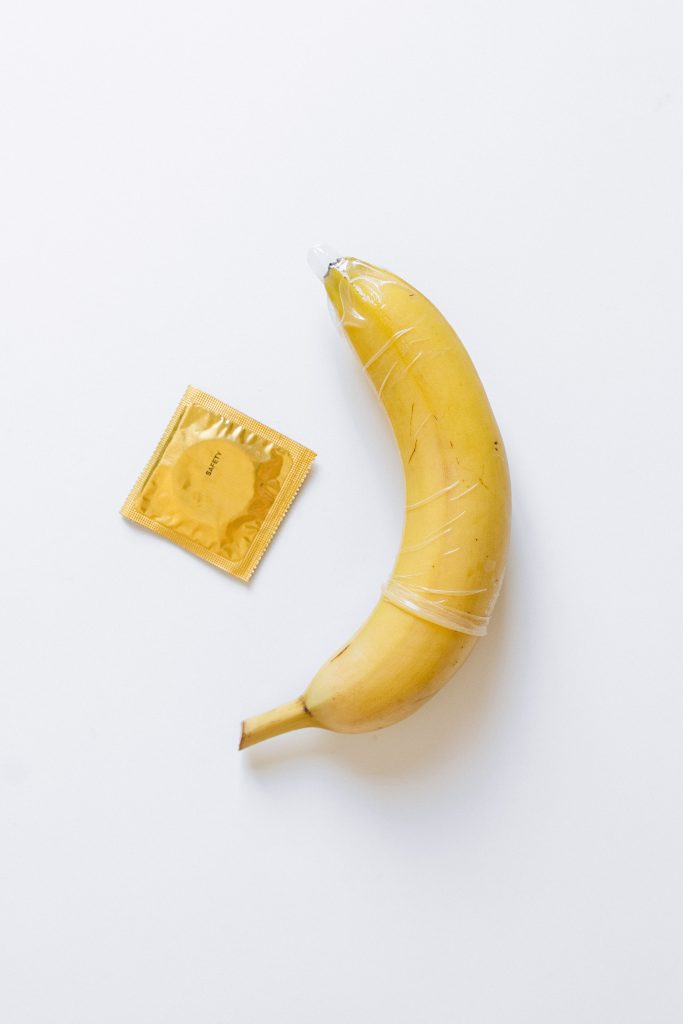 A condom on a banana next to a condom in a gold wrapper.