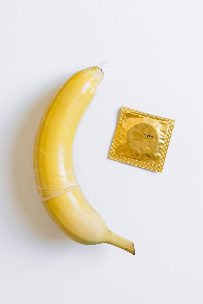 A banana with a condom on it. There is a gold condom in its wrapper beside the banana.