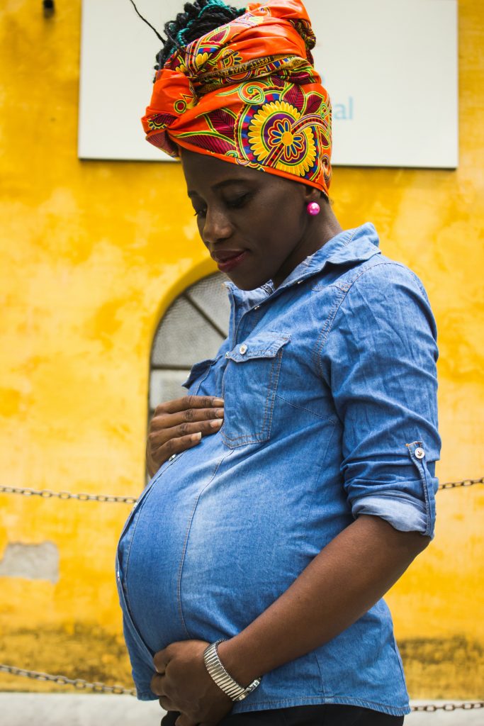 A pregnant person holding their stomach wearing a denim shirt and orange head scarf. 