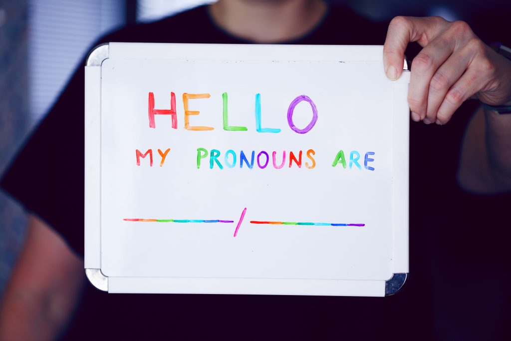 A person holds a white board with "hello my pronouns are blank slash blank" written on it in rainbow colors